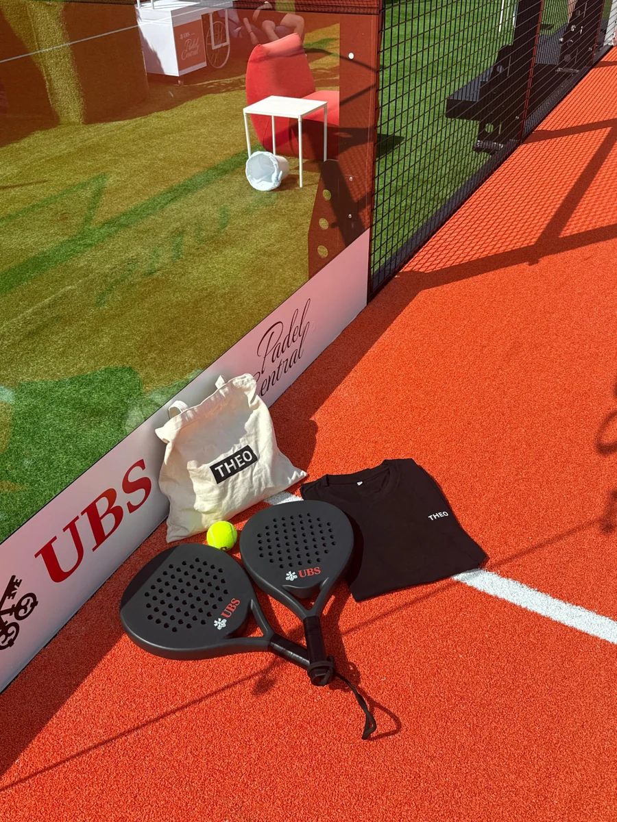 THEO tote bag and t-shirt beside UBS-branded padel rackets and a ball courtside at the UBS Padel Central court.